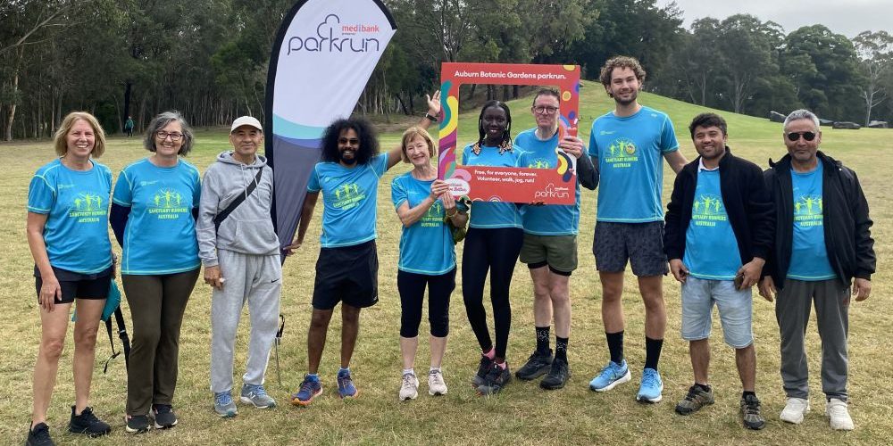 A group of people stand with a Parkrun banner and Auburn Botanic Parkrun branded photo frame in front of a grassy hill, wearing blue Sanctuary Runners Australia tshirts