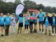 A group of people stand with a Parkrun banner and Auburn Botanic Parkrun branded photo frame in front of a grassy hill, wearing blue Sanctuary Runners Australia tshirts